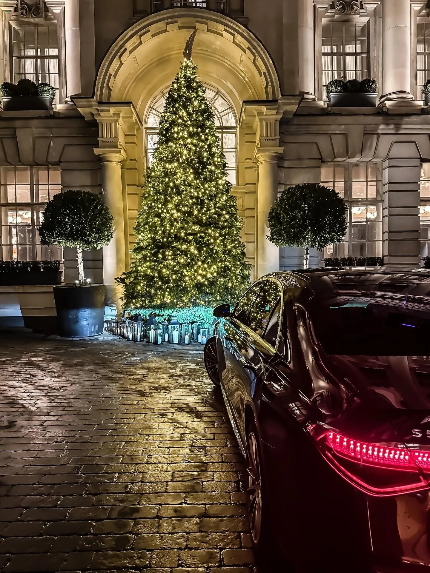 Rear light of a black Mercedes S-Class outside a grand hotel entrance with a lit Christmas tree at night